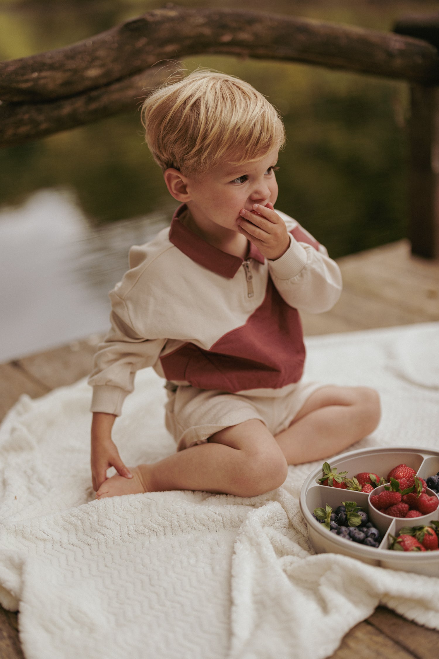 Jongen in rode Viggo sweater van jongensmerk Mr. Loutre geniet van wat fruit terwijl hij op de vlonder zit